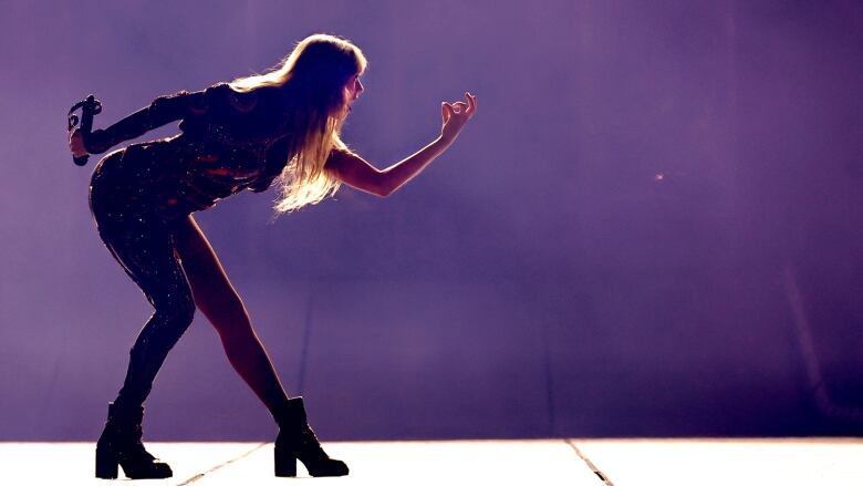 Taylor Swift backlit on stage and gesturing to someone not pictured, while performing.