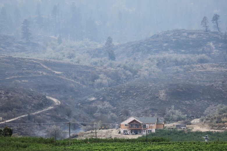 A blackened hillside is visible behind a new home under construction. Grey smoke lingers in the air.