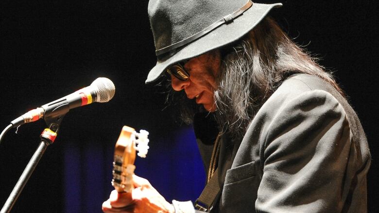 A man with long hair and sunglasses bends over a guitar he is playing. 