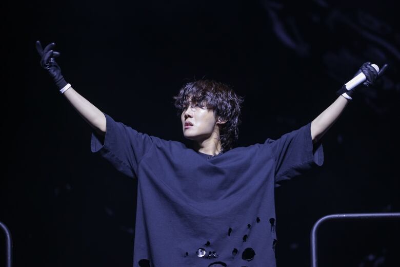 Singer in a baggy black shirt gestures while on stage, holding a microphone in one hand