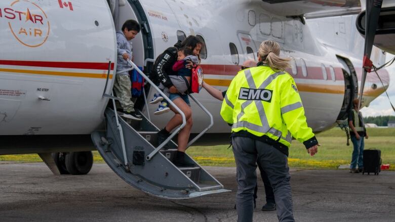 A woman carrying a toddler and a boy disembark a plan as an EMO worker wearing a bright yellow jacket stands next to them.