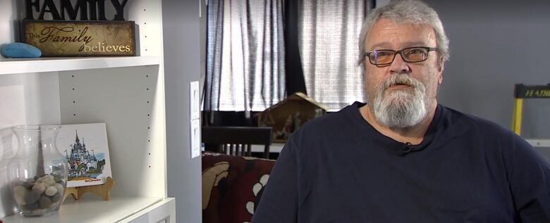 Man with grey hair and beard, wearing glasses and a blue shirt, sits beside a shelf with vase, artwork.