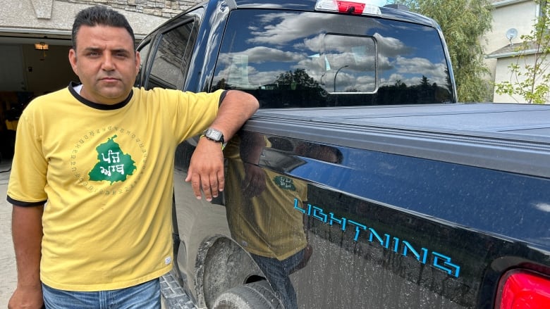 A man in a yellow shirt and blue jeans stands beside the box of a black pickup truck using his left arm to lean on the vehicle.