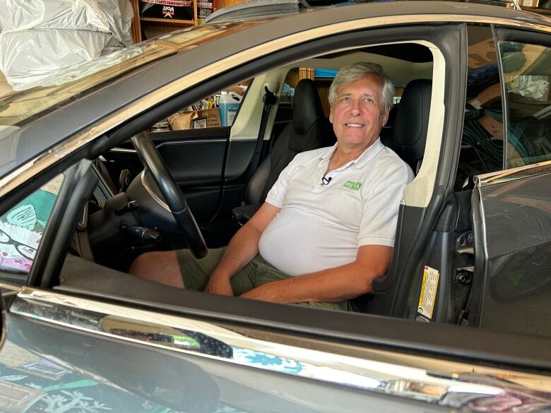 A man in a white shirt sits in the driver's seat of a car with the driver's door cracked halfway open.