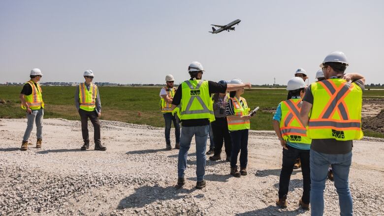 A group of people in orange hi-viz vests and yellow hard hats stands on a gravel field on a sunny day. A plane is seen in the background as it takes off.