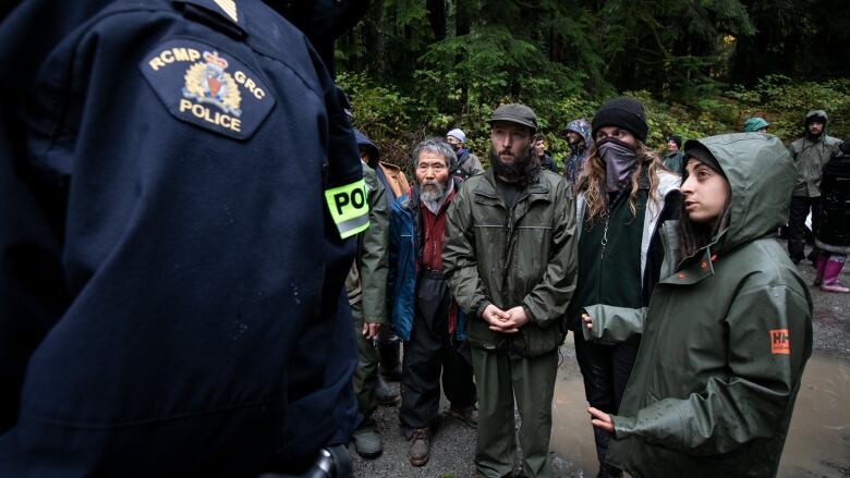 A group of rain-soaked protesters stand on a gravel forest service road looking toward an RCMP officer in a dark blue uniform.