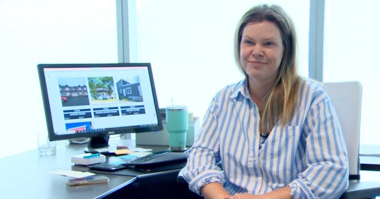 A woman with long hair in a blue and white striped shirt sits at a desk in front of a computer.