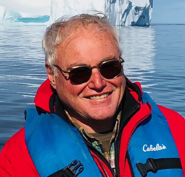 A smiling man in a life jacket and sunglasses is seen with water and an iceberg behind him.