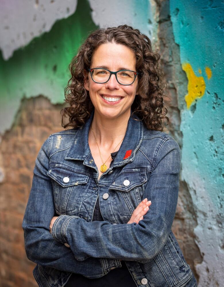 A woman with curly hair and glasses smiles at the camera. The background is blurred.