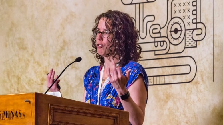 A woman with curly hair and glasses stands at a podium with a microphone and delivers a talk.