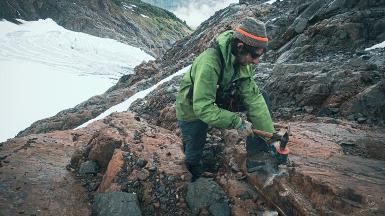 A man with a toque, sunglasses and green jacket uses an excavation tool to take rock samples on snow-covered mountainside.