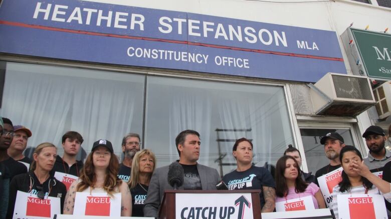 A man is shown speaking at a podium, with people all around him. Many hold picket signs. Above them, a sign saying 