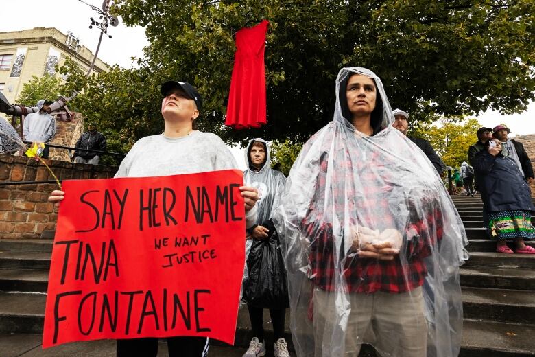 People stand on stairs in front of a tree. One person holds a red sign that says 