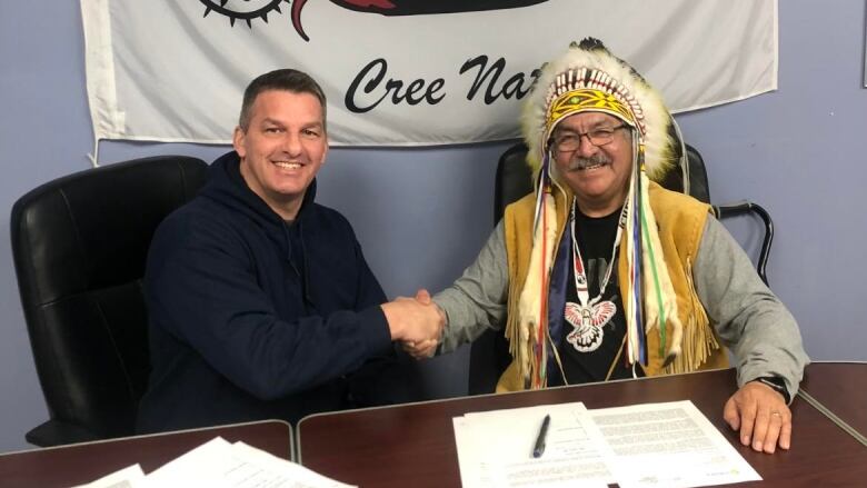 two men sitting at desk, shaking hands and smiling at the camera