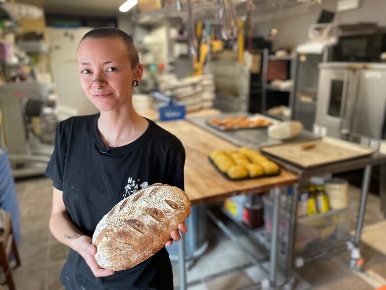 A young person with a buzzcut and several nose piercings smiles and holds a loaf of bread in a bakery.