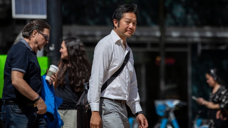 A man with a white open-collared button-up shirt and slacks looks at the camera while walking in front of the B.C. Supreme Court.