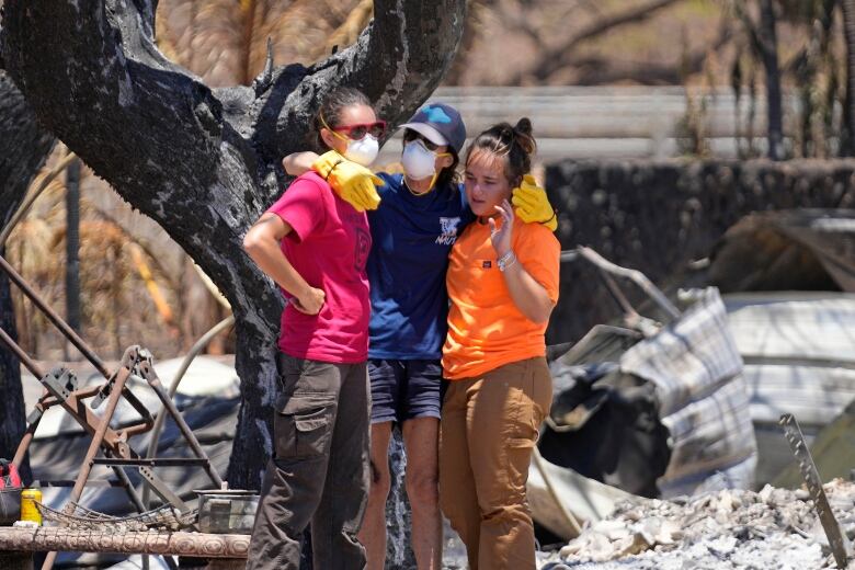 A woman wearing a face mask and yellow gloves puts her arms around the shoulders of two other women, standing amid burned metal and an ashen tree.