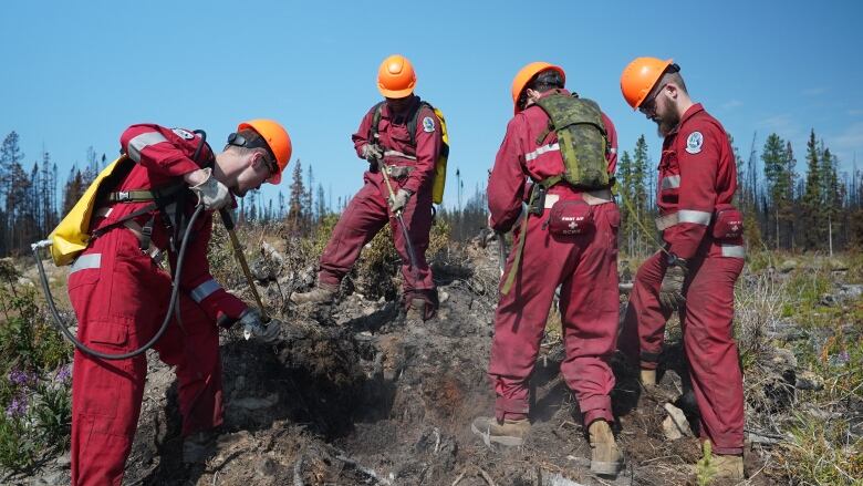 Four men wearing red uniforms poke at smouldering ground with metal instruments.