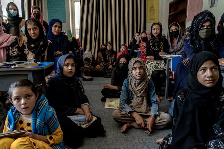 A large group of girls wearing headscarves sit at desks and on the floor in a makeshift classroom.