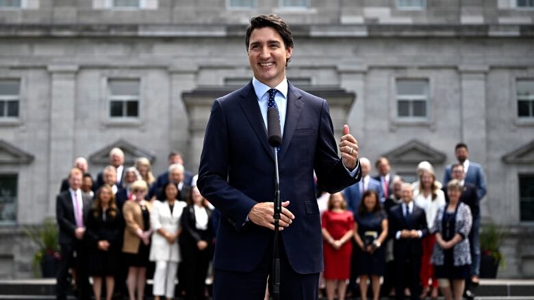 Prime Minister Justin Trudeau stands at a microhpone in front of Rideau Hall, a gray stone building. He is wearing a dark suit and tie. His cabinet stands in the background behind him.