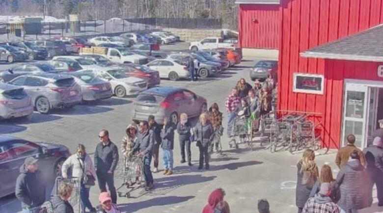 A long lineup of patrons wait to enter a red grocery store. 