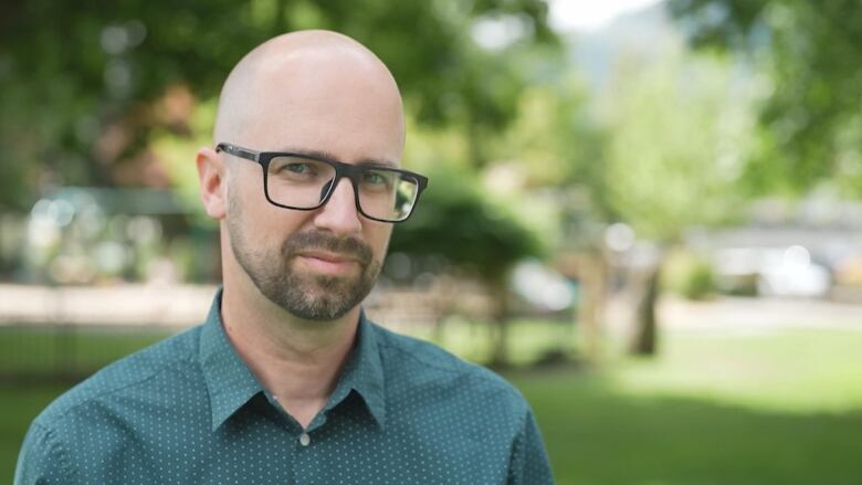 A bald man with a beard looks into the camera with trees behind him