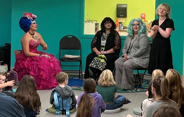 Three drag queens are pictured sitting on chairs in front of a group of children seated on the floor.