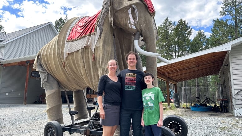 A man, a woman and a boy pose in front of a giant elephant sculpture on wheels that dwarfs them.