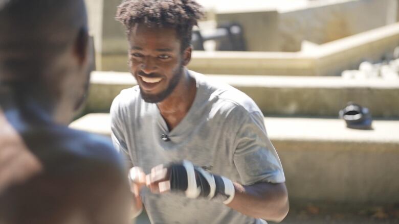 A 20-year-old man spars with wraps around his two hands and sweat on his T-shirt in a training session. One of his trainers, blurred in the foreground, faces him.