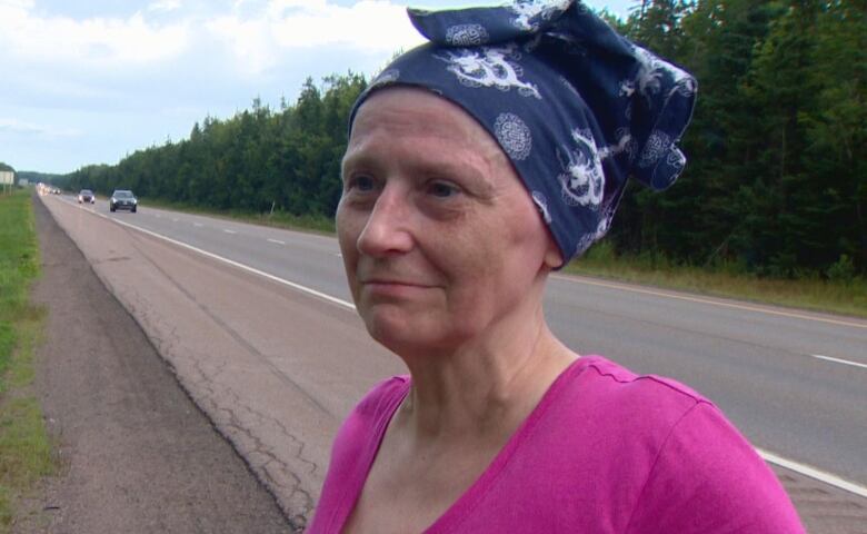 A woman in a pink shirt standing on the side of the road. Her hair is covered by navy blue and white head covering.