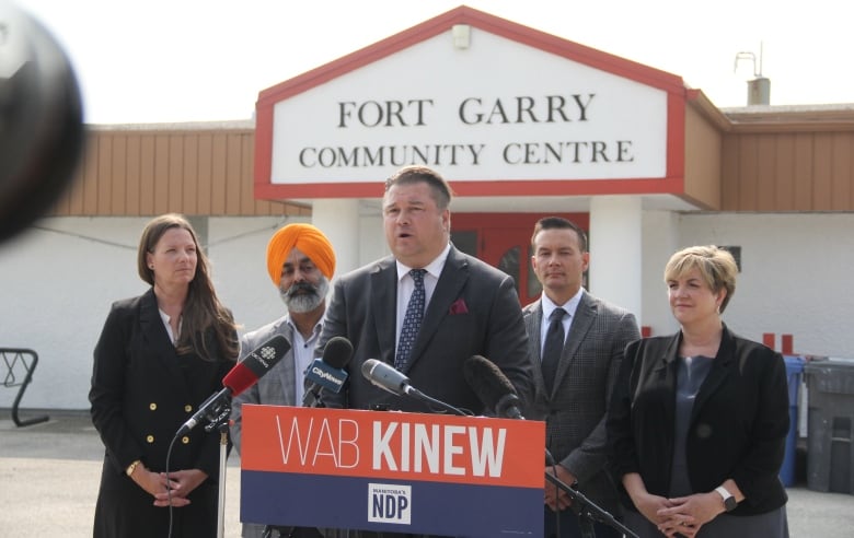 Several politicians stand behind a podium, in front of the front entrance of a community centre.