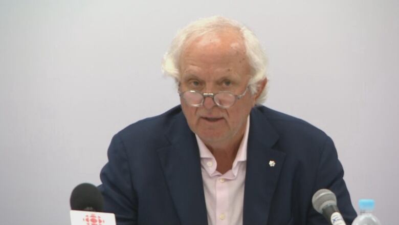 Man in navy blue blazer and pink button-up shirt sits in front of multiple mics with grey backdrop.