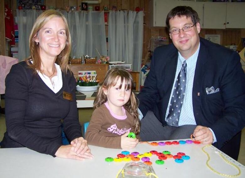 A woman, left, a small girl playing with a toy, middle, and a man in a suit, right.