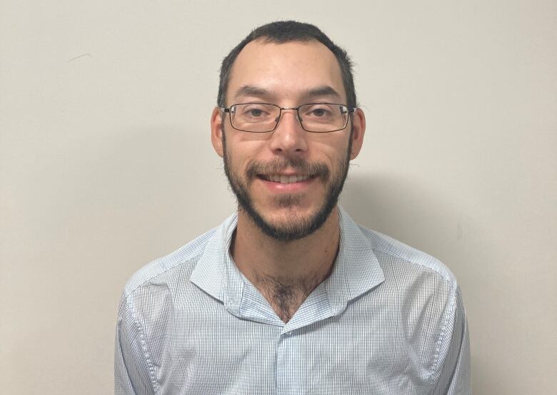 A smiling man in a baby blue dress shirt in front of a cream-coloured wall.