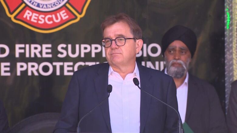 A white man wearing glasses delivers a news conference, with a turbaned man behind him.