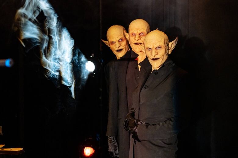 Medium shot of three performers wearing identical goblin masks, standing together on a darkened stage.