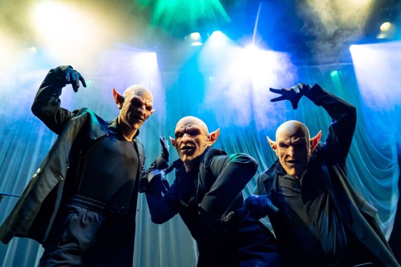 Medium shot, photographed from below. Three performers in black wearing realistic goblin masks. They strike dramatic poses, raising their arms above their heads, on a stage draped with blue curtains.