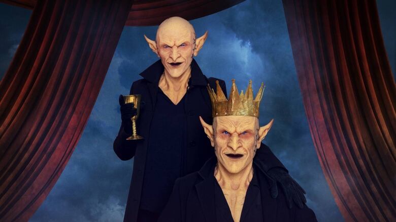 Portrait of two performers wearing realistic goblin masks. One is seated and wears a crown. The other stands next to the seated goblin, holding a brass goblet. The background is deep blue and the figures are flanked by burgundy drapes.