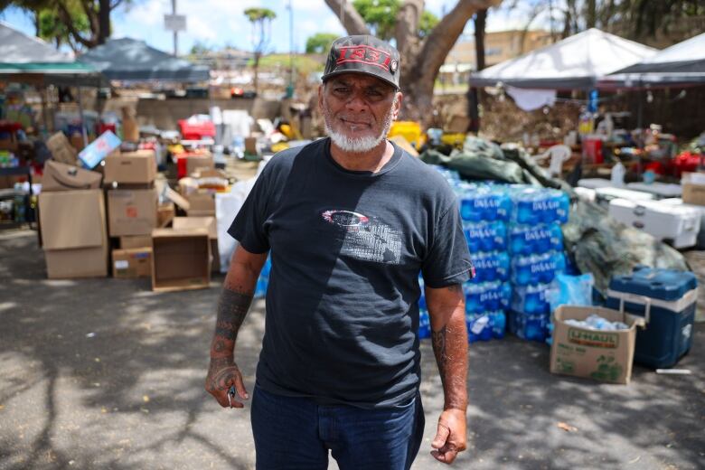 Keeaumoku Kapu stands in front of pop up tents and piles of suplies in a parking lot. He wears jeans, a black t-shirt and cap with red writing. 