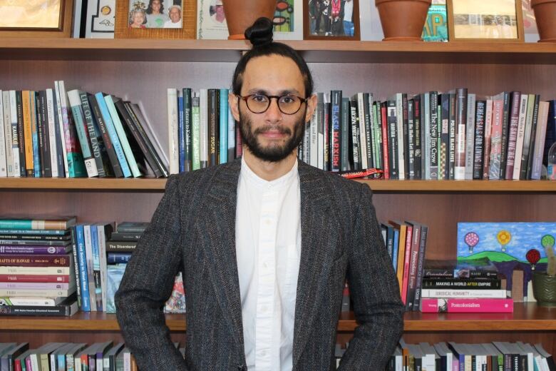 Uahikea Maile stands in front of a book case looking at the camera in white button up shirt, and grey pin stripped blazer. He has thick framed round glasses and black hair in a bun on top of his head. 