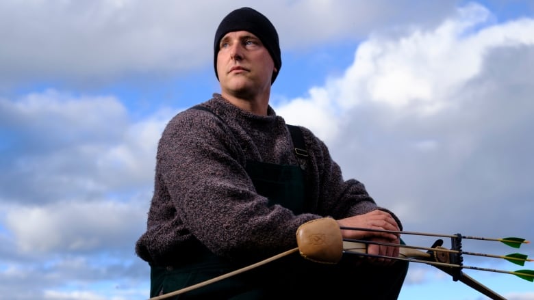 A man crouches in front of an ocean, holding a bow and arrows. 