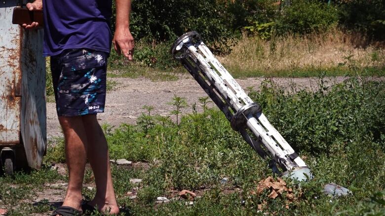  A man, cropped from the torso down, walks past a spent rocket stuck in the grass-covered ground with a house in the background.