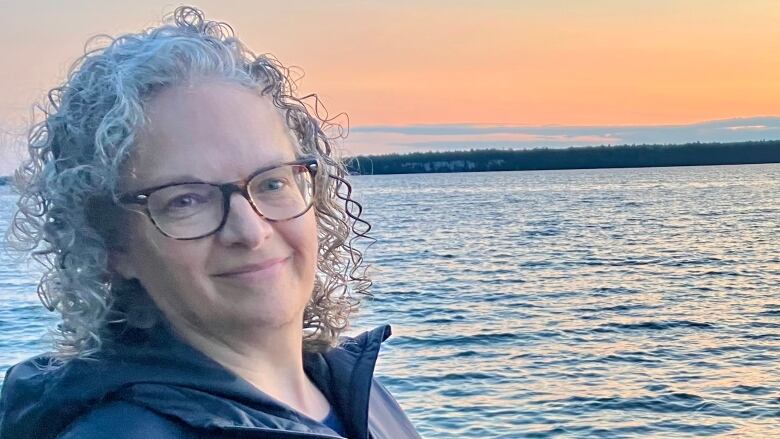 a woman wearing glasses, with curly hair, smiling in front of a water body