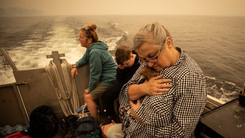 A middle-aged woman with grey hair cradles a small brown dog inside her black and white plaid shirt as she rides in an aluminum boat on a lake.