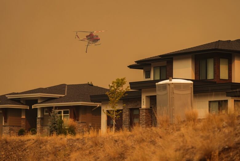 A helicopter flies through brown smoky skies, above a row of large, modern homes.