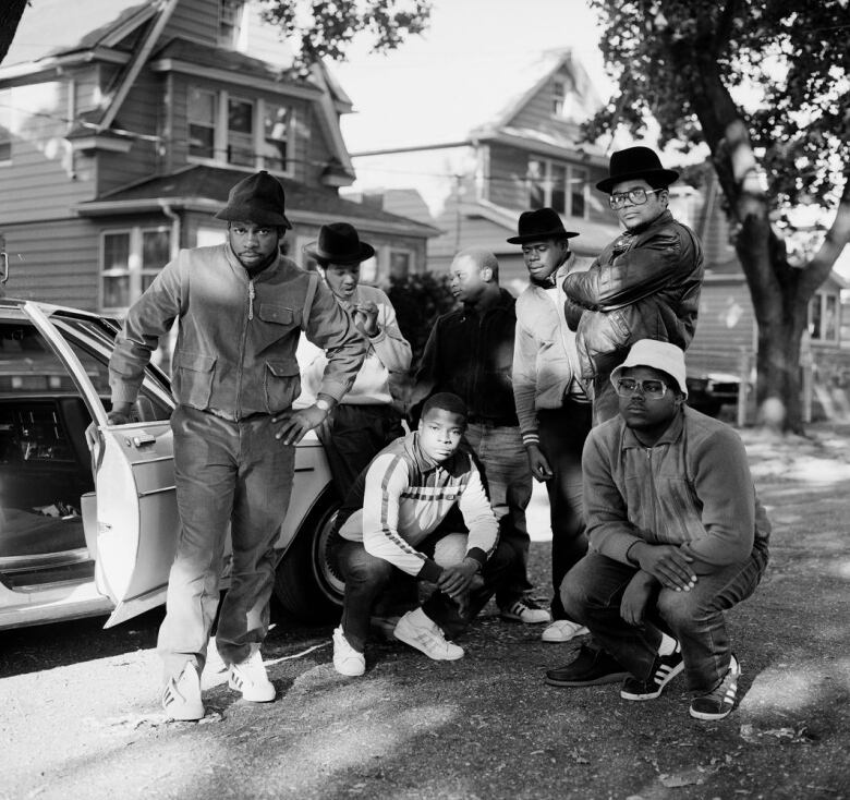 Black and white group portrait of Run-D.M.C. and three members of their posse standing outside in a residential neighbourhood.