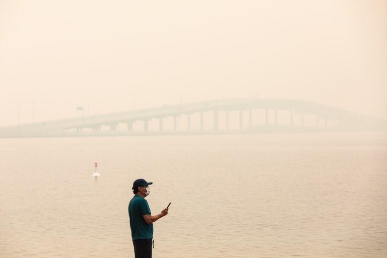 A man in a green T-shirt, black shorts, black baseball hat and white mask stands at the edge of a lake. The air is thick with smoke.