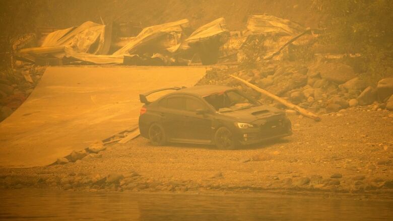 A black sports car, with the front window smashed, sits beside a lake, the air around it is brown and hazy, with fire-destroyed property behind it.