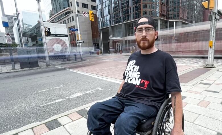 Kyle Humphrey is a man with a red beard, glasses and a backwards cap. He looks into the camera and sits in a wheelchair on a busy downtown street. His shirt reads 