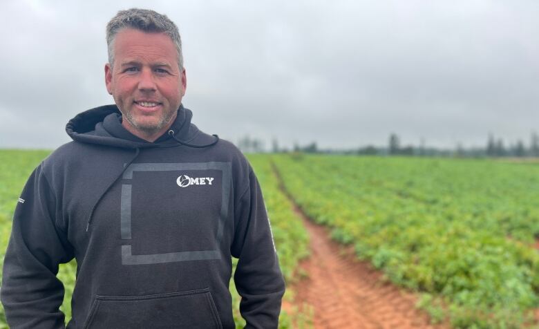 A man stands in a potato field smiling and wearing a black hoodie and jeans. 